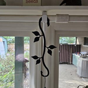 May include: Black metal decorative vine with leaves, hanging from a white clip on a window frame. The vine has a hook at the bottom, possibly for hanging a wind chime or other small item. The background shows a window and outdoor scenery.