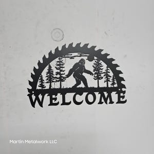May include: Black metal welcome sign featuring a Bigfoot silhouette walking through a forest scene. The sign is framed by a saw blade design and includes the word "WELCOME" at the bottom. The text "Martin Metalwork LLC" is at the bottom.