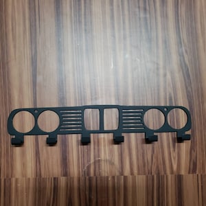 May include: Black metal wall-mounted coat rack with a car grill design. Features two circular openings on each end, a rectangular opening in the center, and horizontal slats. Includes four hooks for hanging items.
