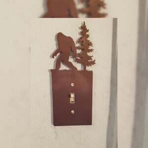 May include: A brown metal light switch cover featuring a Bigfoot silhouette walking towards a tree. The switch cover is rectangular with a toggle switch and two screws. The Bigfoot and tree are cut-out designs.