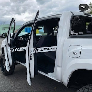 May include: White pickup truck with open doors, showcasing custom door bars. The bars are white with black text that reads "TACCO" and "SUPREME". The truck has black wheels and tinted windows. The image is taken outdoors.