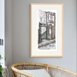 Op de afbeelding: Een ingelijste aquarelprint van The Cavern Club, een historische muzieklocatie. Het kunstwerk toont de buitenkant van de club met bewegwijzering, metselwerk en een open deur. De tekst op de print luidt "The Cavern" en "The Original Cavern Club 1963."
