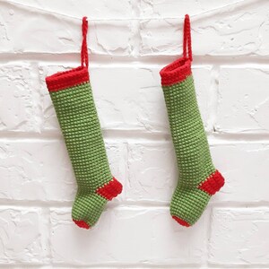 Op de afbeelding: Twee groene en rode gehaakte kerstsokken die aan een witte bakstenen muur hangen.