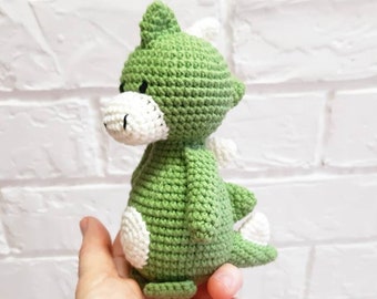 May include: A handmade green and white crochet dinosaur toy. The amigurumi dinosaur has a green body with white accents on its belly, feet, and wings. The toy is held in a hand against a white brick wall.