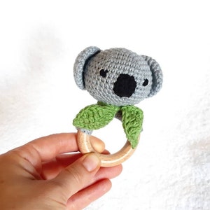 May include: A handmade crochet baby rattle featuring a gray koala head with black eyes and nose, and green leaves. The koala is attached to a natural wood ring. A soft, safe toy for infants.