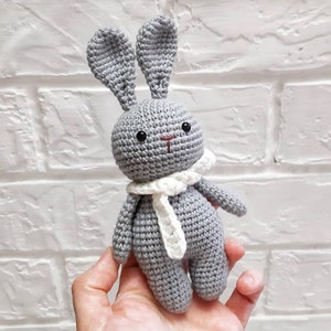 Puede incluir: Un conejito de crochet gris con orejas y una bufanda de crochet blanco. El conejito est&aacute; sentado en la mano de una persona.