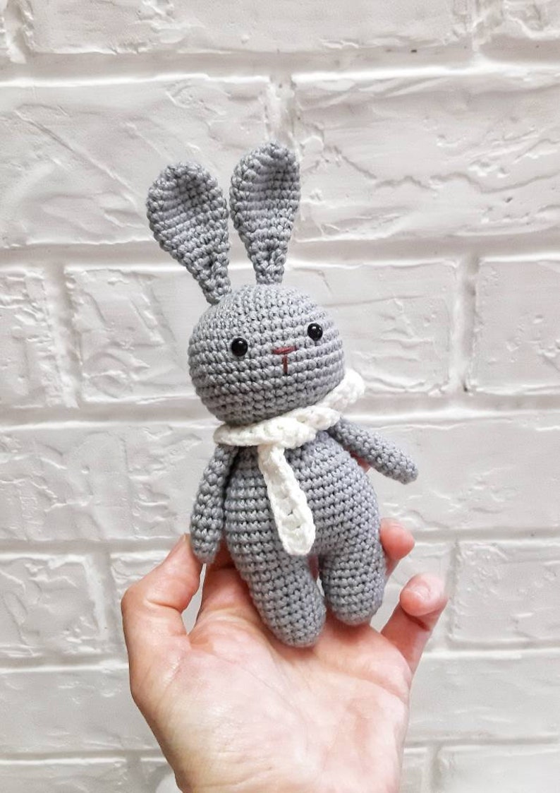 Puede incluir: Un conejito de crochet gris con orejas y una bufanda de crochet blanco. El conejito est&aacute; sentado en la mano de una persona.