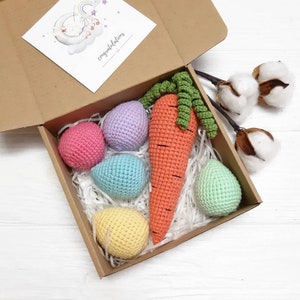 Puede incluir: Una caja de regalo marr&oacute;n con relleno de papel de seda blanco. La caja contiene una zanahoria de ganchillo y cinco huevos de ganchillo en varios colores pastel. La caja est&aacute; abierta y la tapa est&aacute; en el lateral. El texto "felicitaciones" est&aacute; escrito en la tapa.