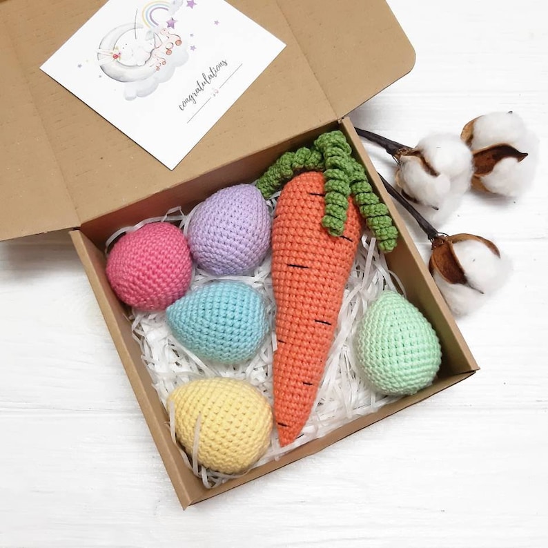 Puede incluir: Una caja de regalo marr&oacute;n con relleno de papel de seda blanco. La caja contiene una zanahoria de ganchillo y cinco huevos de ganchillo en varios colores pastel. La caja est&aacute; abierta y la tapa est&aacute; en el lateral. El texto "felicitaciones" est&aacute; escrito en la tapa.