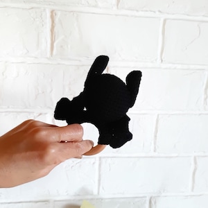 May include: Black crochet baby rattle in the shape of a bat with a wooden ring. The rattle is held against a white brick wall. The bat has a round head, two pointed ears, and outstretched wings.