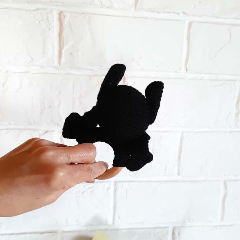 May include: Black crochet baby rattle in the shape of a bat with a wooden ring. The rattle is held against a white brick wall. The bat has a round head, two pointed ears, and outstretched wings.