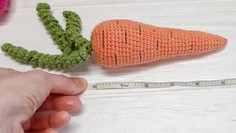 May include: A crocheted carrot toy with green leaves, measuring 12.7 cm long. The carrot is orange with black lines and the leaves are green.