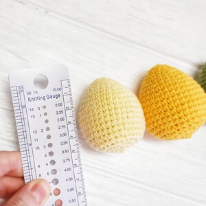 May include: A close-up of a knitting gauge with a yellow, light yellow, and green crocheted egg beside it. The gauge is marked with numbers and dots to indicate different knitting needle sizes.