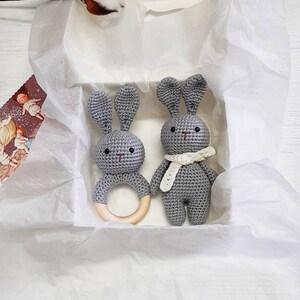 Puede incluir: Dos juguetes de conejo de crochet gris con anillos de dentici&oacute;n de madera. Un conejo lleva una bufanda de crochet blanca.