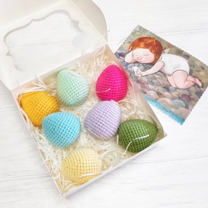 May include: A white gift box filled with colorful crocheted Easter eggs. The eggs are in shades of yellow, green, blue, pink, and purple. The box is lined with white shredded paper.