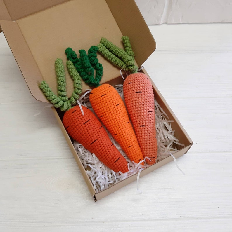 May include: Three crocheted carrots in a brown cardboard box with white shredded paper. The carrots are orange with green tops.