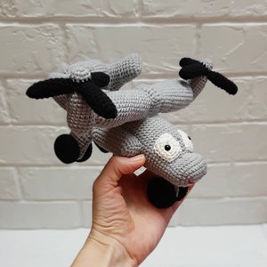 Può includere: Un giocattolo elicottero in crochet grigio con eliche nere e occhi bianchi. Il giocattolo è tenuto in una mano.