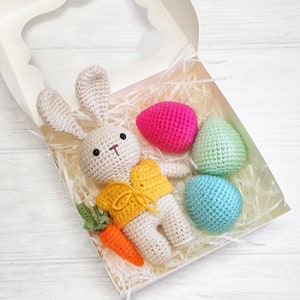 May include: A crocheted bunny wearing a yellow vest and holding a carrot is in a white gift box with three crocheted Easter eggs in pink, green, and blue.