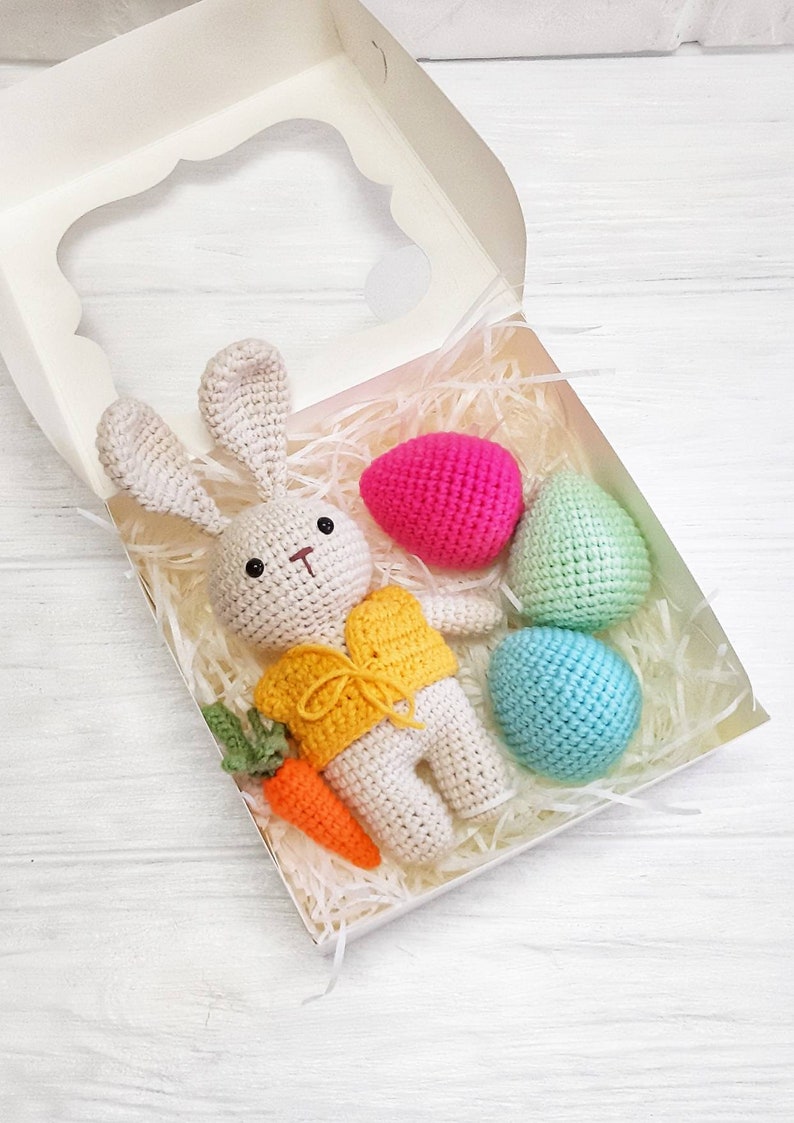 May include: A crocheted bunny wearing a yellow vest and holding a carrot is in a white gift box with three crocheted Easter eggs in pink, green, and blue.