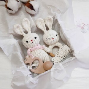 Puede incluir: Dos juguetes de conejo de crochet blanco con detalles en rosa y blanco. Un conejo tiene un mordedor de anillo de madera y el otro tiene un mordedor de madera con forma de corona. Los juguetes est&aacute;n en una caja de regalo blanca con papel de seda.