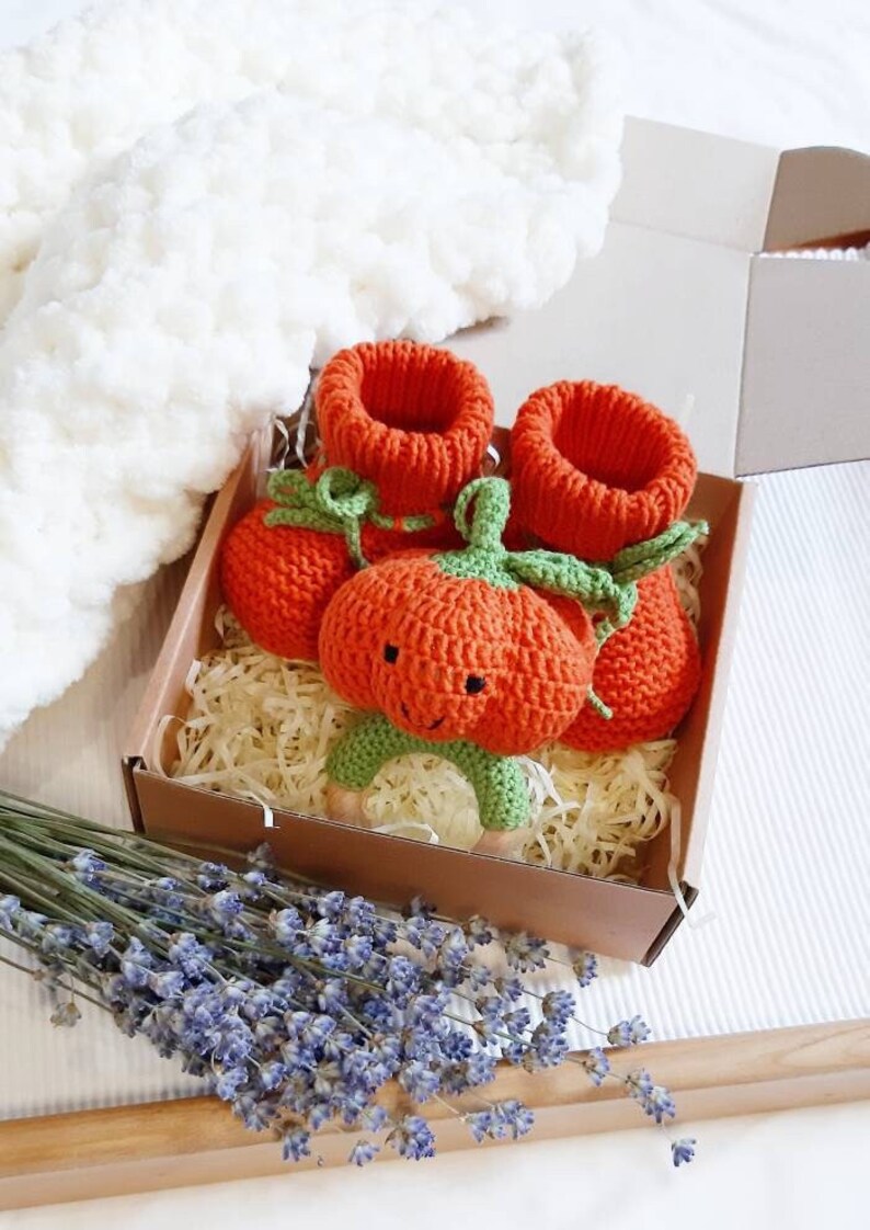 May include: A set of orange knitted pumpkin baby booties with green leaves and a matching orange knitted pumpkin rattle with a smiling face and green leaves. The set is packaged in a brown box with shredded paper.