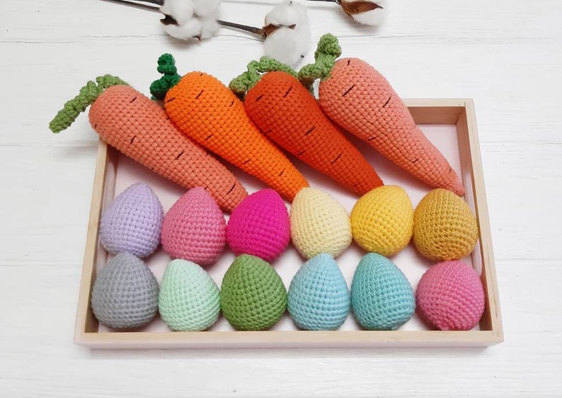 May include: A wooden tray filled with crocheted carrots and eggs in various pastel colours. The carrots are orange and green, and the eggs are lavender, pink, red, yellow, light green, blue, and light pink.