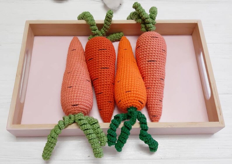 May include: Four crocheted carrots in a wooden tray with a pink liner. The carrots are orange and pink with green tops.