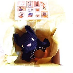 May include: A black crochet bat-shaped baby rattle with a wooden ring, presented in a box with yellow tissue paper. A card with Halloween-themed stickers is visible. The rattle has white eyes and a pink mouth.