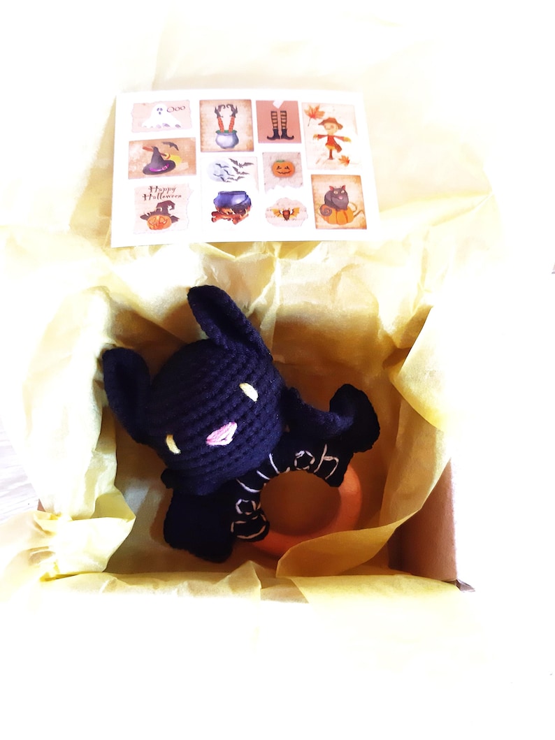 May include: A black crochet bat-shaped baby rattle with a wooden ring, presented in a box with yellow tissue paper. A card with Halloween-themed stickers is visible. The rattle has white eyes and a pink mouth.