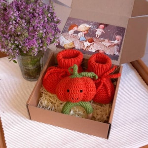 May include: A brown cardboard box with a red knitted baby bootie, a red knitted tomato rattle, and a green and brown wooden ring rattle. The box is filled with shredded paper.