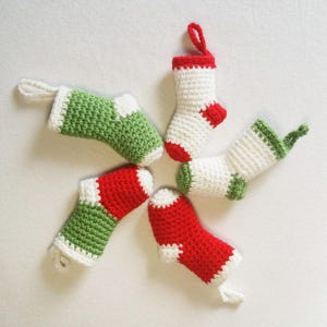 Op de afbeelding: Een verzameling van vijf miniatuur gehaakte kerstsokken in een stervormig patroon. De sokken zijn rood, wit en groen, met witte manchetten en ophanglussen. De handgemaakte ornamenten zijn een feestelijke vakantieversiering.