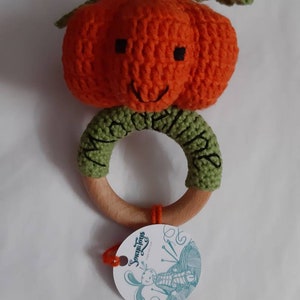 May include: A crocheted pumpkin rattle with a wooden ring and a green stem. The pumpkin has a smiling face and is attached to the wooden ring with green crocheted fabric. The tag says "Something for Little Ones".