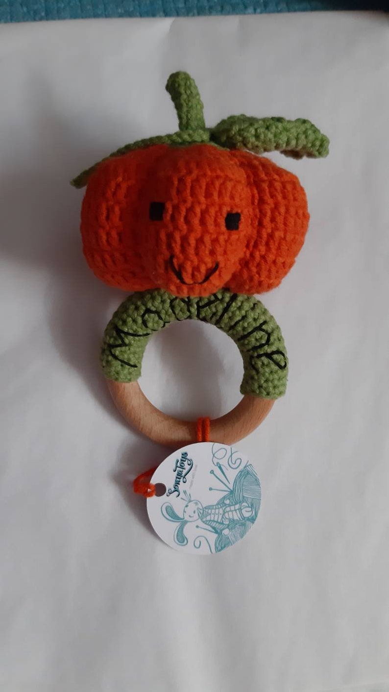 May include: A crocheted pumpkin rattle with a wooden ring and a green stem. The pumpkin has a smiling face and is attached to the wooden ring with green crocheted fabric. The tag says "Something for Little Ones".