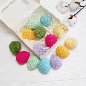 May include: A collection of crocheted Easter eggs in a variety of colors, including pink, green, yellow, blue, and purple. The eggs are arranged in a white box with shredded paper.