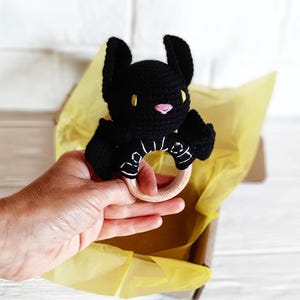 May include: A black crochet bat-shaped baby rattle with a wooden ring. The bat has yellow eyes and a pink nose. The rattle is held in a hand, inside a cardboard box with yellow tissue paper.