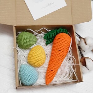May include: A gift box filled with white shredded paper and crocheted Easter decorations. The decorations include three eggs in green, yellow, and blue, and a carrot with green leaves.