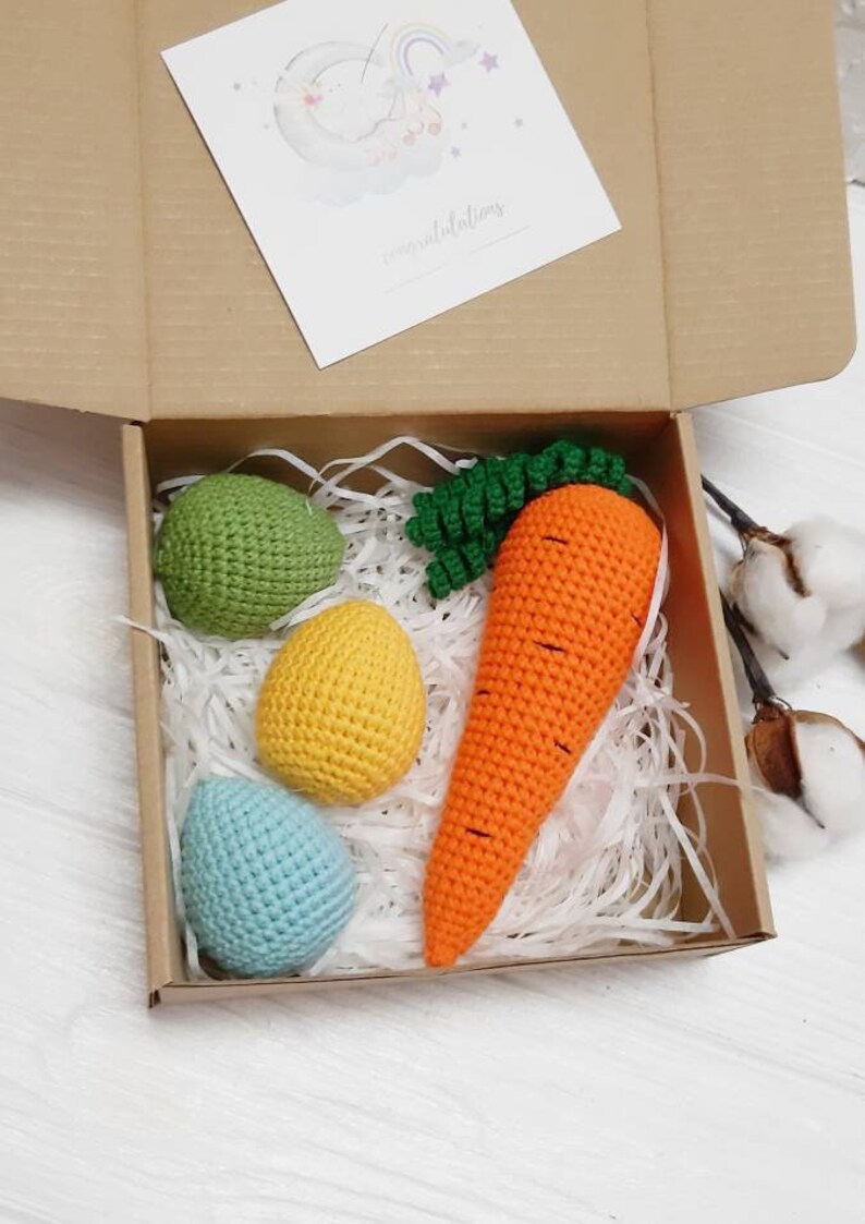 May include: A gift box filled with white shredded paper and crocheted Easter decorations. The decorations include three eggs in green, yellow, and blue, and a carrot with green leaves.