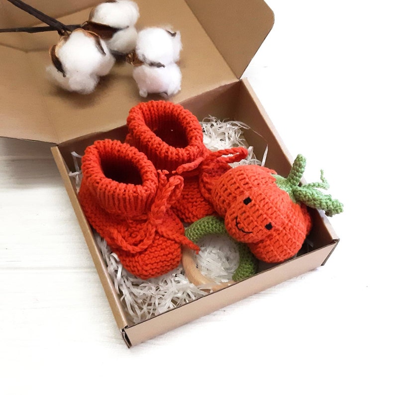 May include: A gift box filled with orange knitted baby booties and a crocheted orange pumpkin rattle with a green stem and a smiling face.