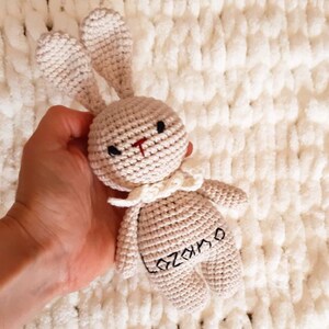 Puede incluir: Un juguete de conejo de crochet beige con un cuello blanco y el nombre "Ozamo" bordado en la espalda.