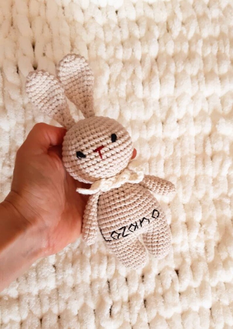 Puede incluir: Un juguete de conejo de crochet beige con un cuello blanco y el nombre "Ozamo" bordado en la espalda.