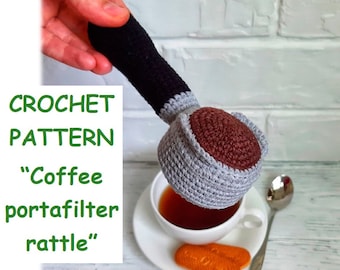 May include: Crochet pattern for a "Coffee portafilter rattle" toy. The rattle is gray and brown and shaped like a coffee portafilter. It is being held over a cup of tea.