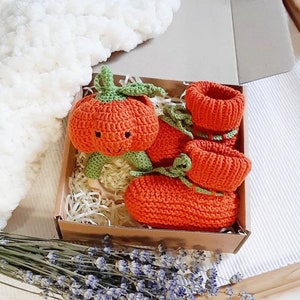 May include: A set of orange knitted baby booties shaped like pumpkins with green stems and a smiling face. The booties are in a brown box with shredded paper.