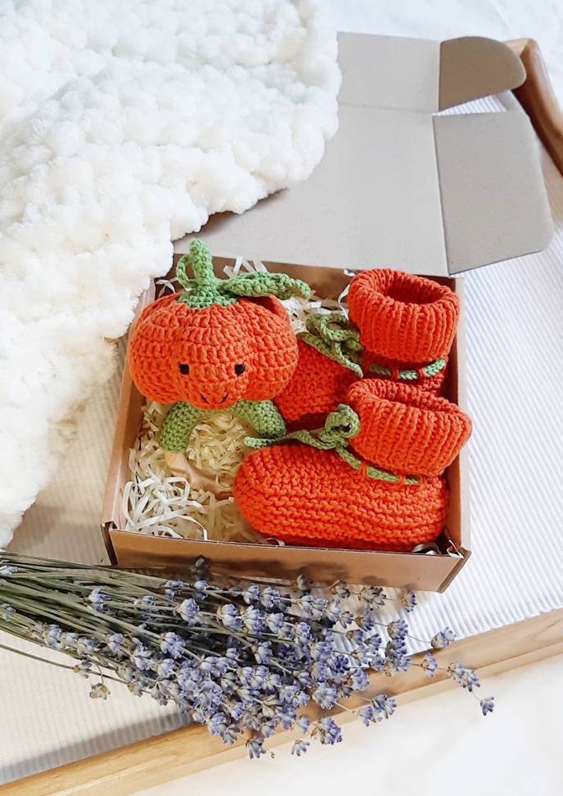 May include: A set of orange knitted baby booties shaped like pumpkins with green stems and a smiling face. The booties are in a brown box with shredded paper.