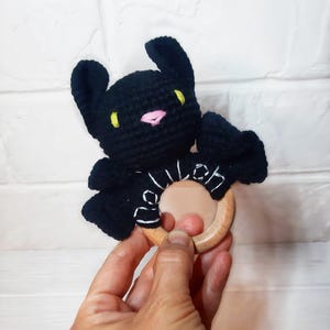 May include: A black crochet bat-shaped baby rattle with yellow eyes and a pink nose. The bat is attached to a natural wood ring. The rattle is held in a person's hand against a white brick background.