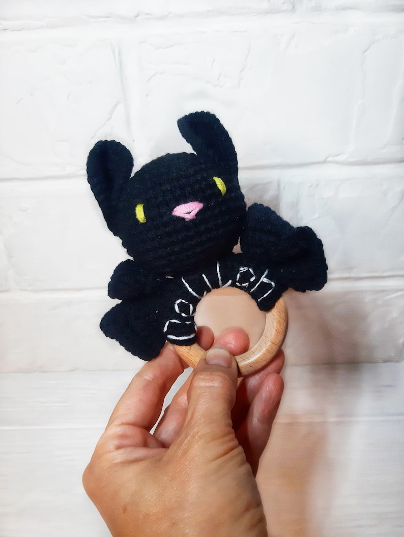 May include: A black crochet bat-shaped baby rattle with yellow eyes and a pink nose. The bat is attached to a natural wood ring. The rattle is held in a person's hand against a white brick background.