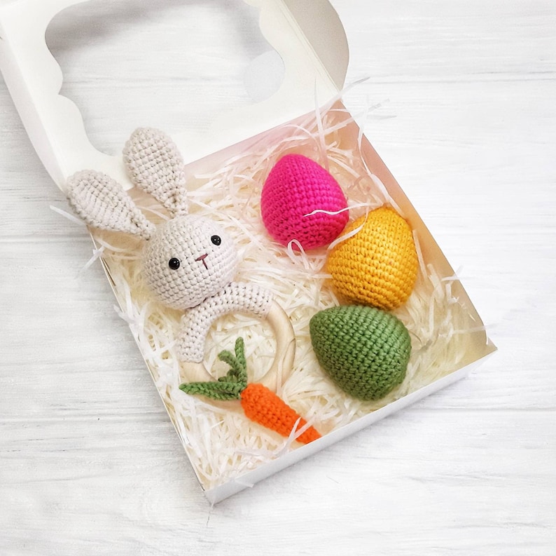 May include: A white crocheted bunny rattle with a wooden ring, a green carrot, and three crocheted Easter eggs in pink, yellow, and green. The items are in a white gift box with shredded paper.