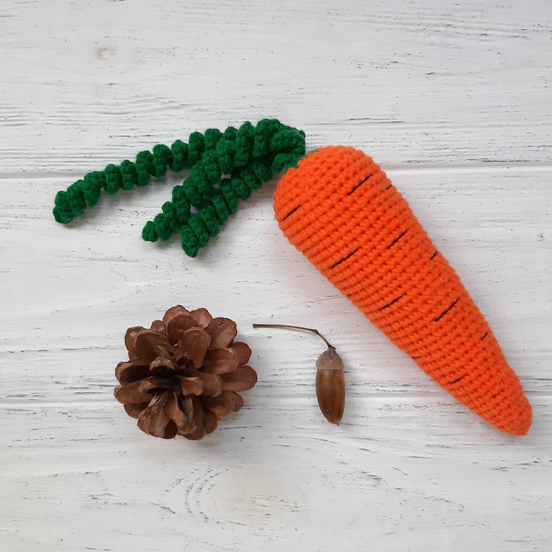 May include: A crocheted carrot toy with green leaves. The carrot is orange with black stitching and is lying on a white wooden surface. There is a brown pine cone and a brown acorn on the surface.