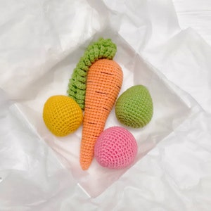 Puede incluir: Un conjunto de juguetes de crochet en una bolsa de regalo de papel de seda blanco. El conjunto incluye una zanahoria, una bola verde, una bola amarilla y una bola rosa.