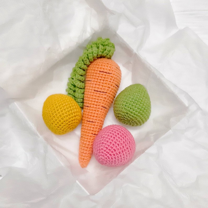 Puede incluir: Un conjunto de juguetes de crochet en una bolsa de regalo de papel de seda blanco. El conjunto incluye una zanahoria, una bola verde, una bola amarilla y una bola rosa.