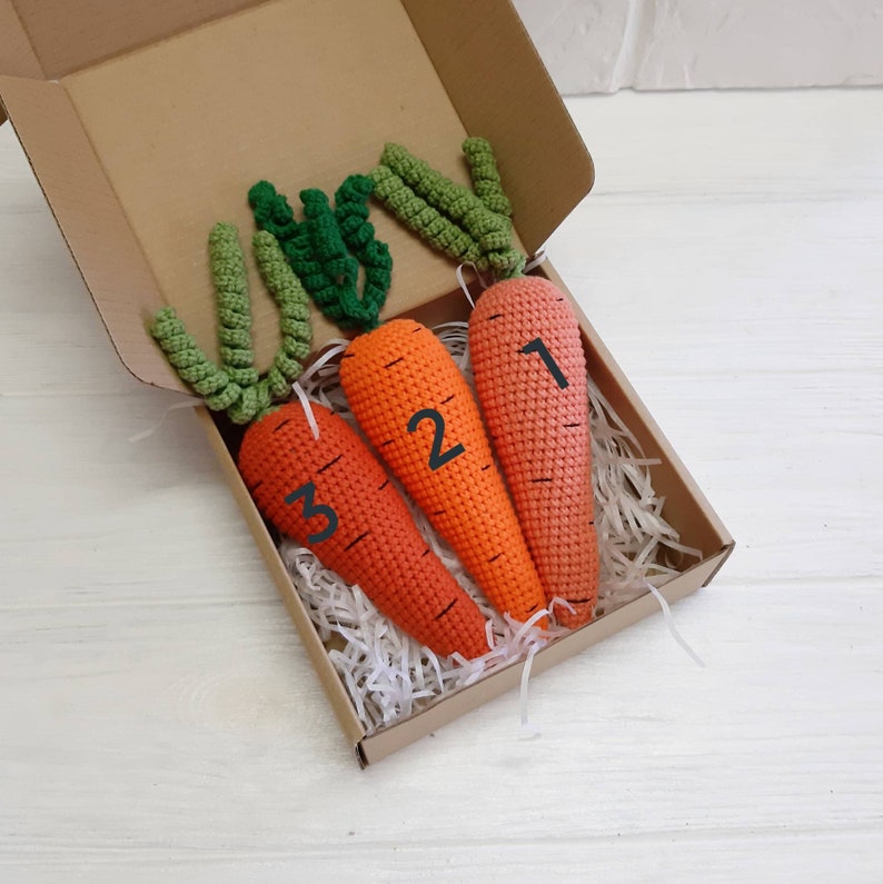 May include: Three crocheted carrots in a box, each with a different number on them. The carrots are orange and green and are arranged in a row.